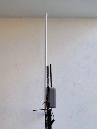 Outdoor antenna installation mounted on a pole against a textured, light gray concrete wall. The setup includes a tall white cylindrical main antenna, a smaller black antenna, and a gray rectangular device (likely a wireless router or modem) attached to the pole, with cables and black zip ties securing the components.